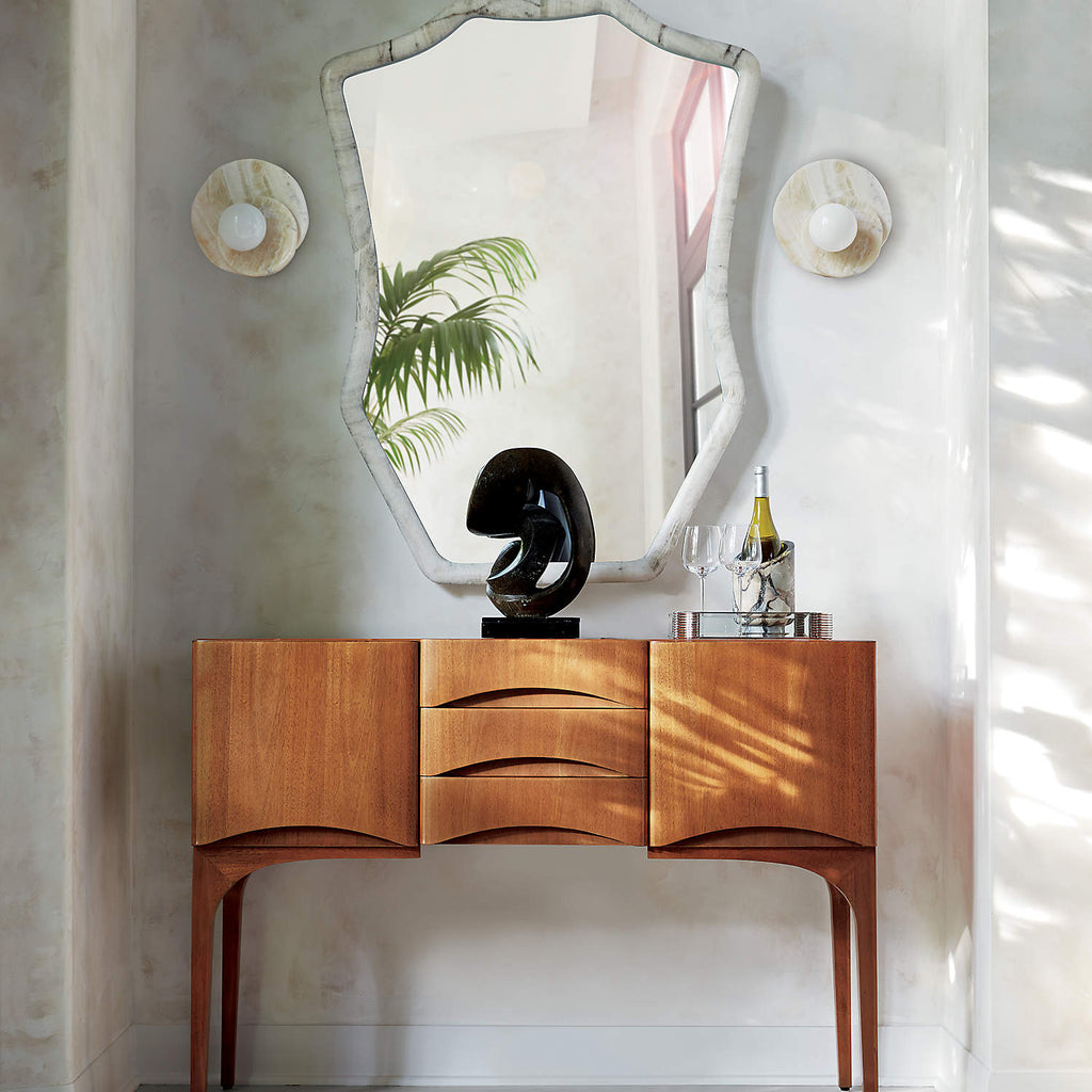 Either side of an arched mirror above a teak sideboard, Clérachic wall lights light a console styled with a small sculpture.