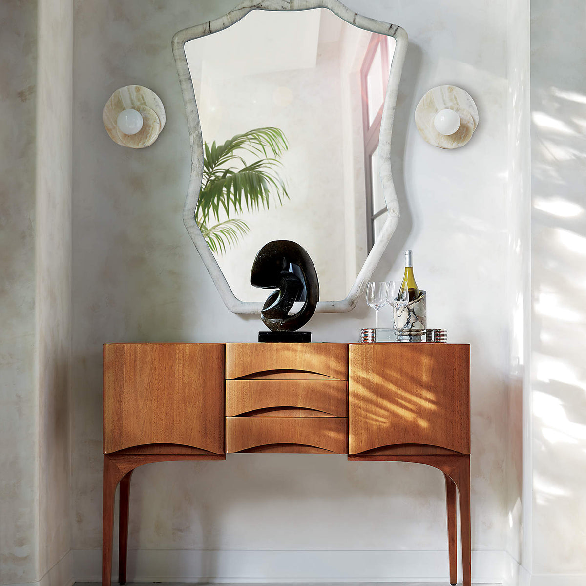 Either side of an arched mirror above a teak sideboard, Clérachic wall lights light a console styled with a small sculpture.