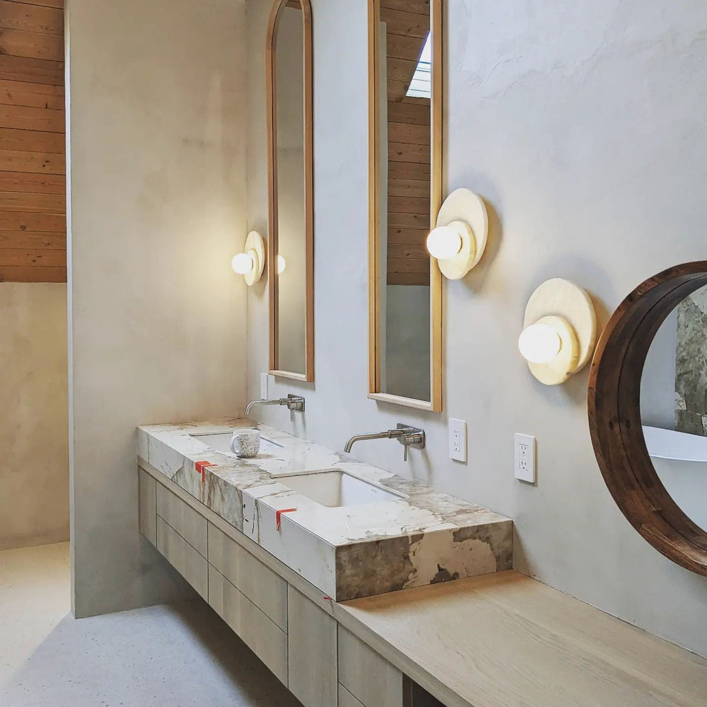 Two Clérachic wall lights flank mirrored vanities in a stone bathroom, lighting a pair of sinks with a soft warm glow.