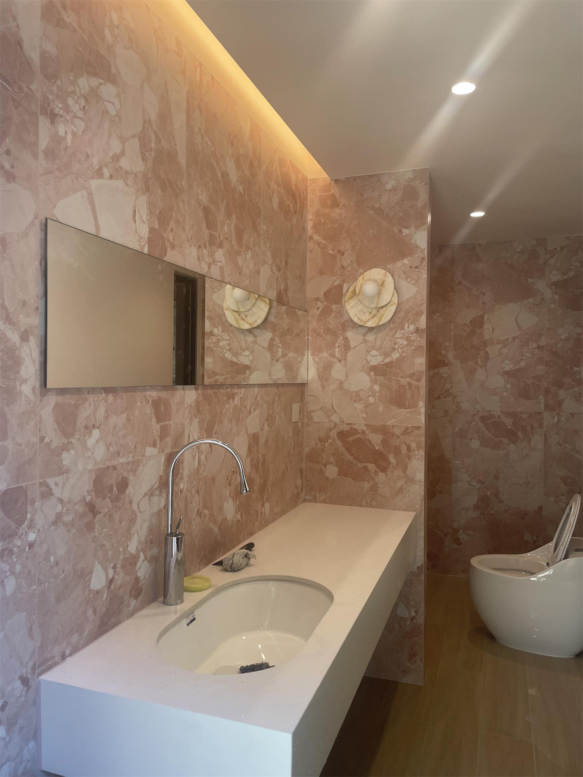 Two Clérachic wall lights flank a mirror in a rose-toned stone bathroom above a long floating vanity with two basins.