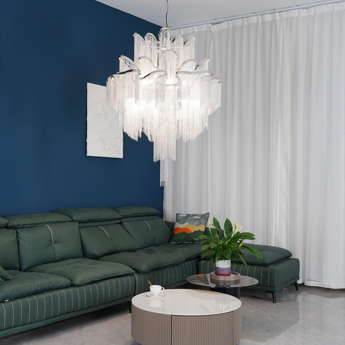 Against a deep blue wall, this lit silver Clérachic chandelier hangs above a green leather sofa and sheer curtains.
