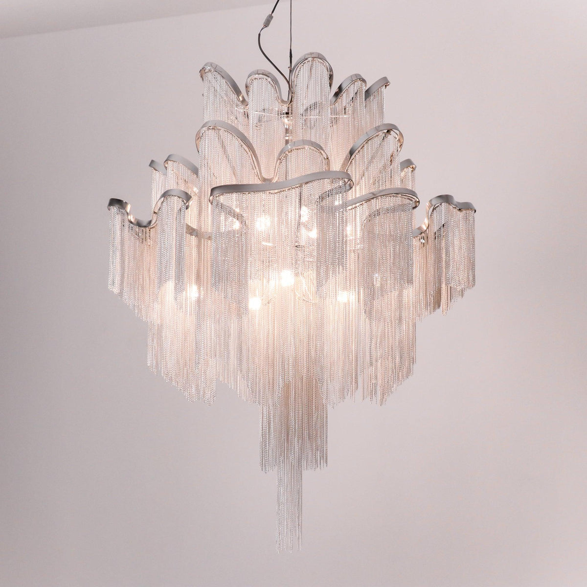 On a soft pink-grey backdrop, this lit silver Clérachic chandelier is shown with its chain tiers glowing.