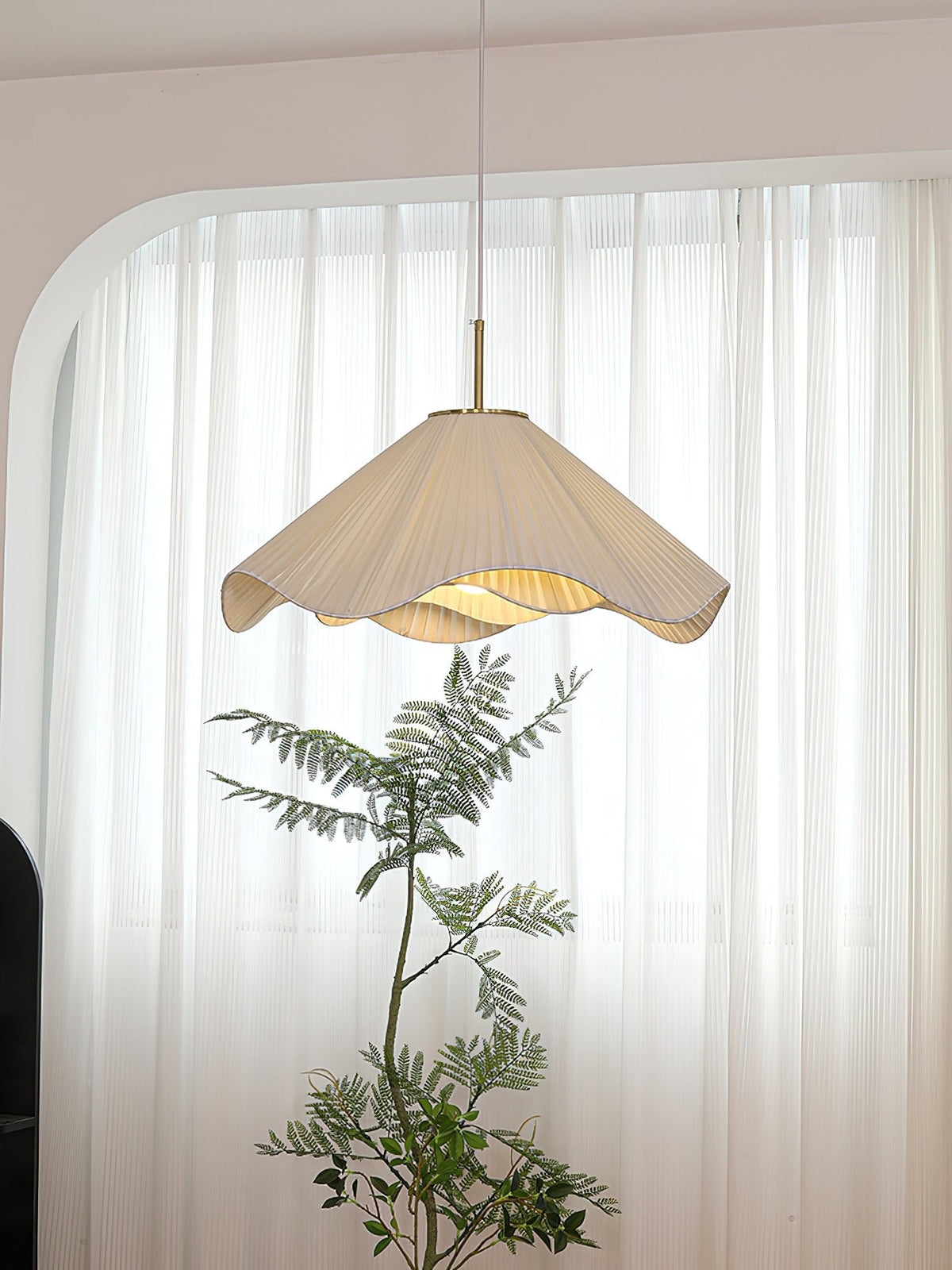 In front of a sheer curtained window, this beige Clérachic pendant light glows beside a tall potted plant.