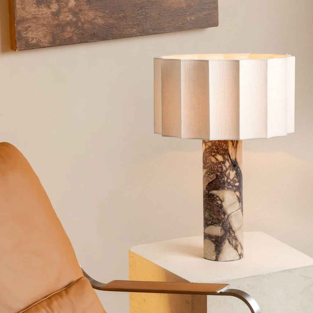 The Clérachic table lamp's faceted cream shade and veined purple marble base stand on a stone plinth beside a tan armchair.