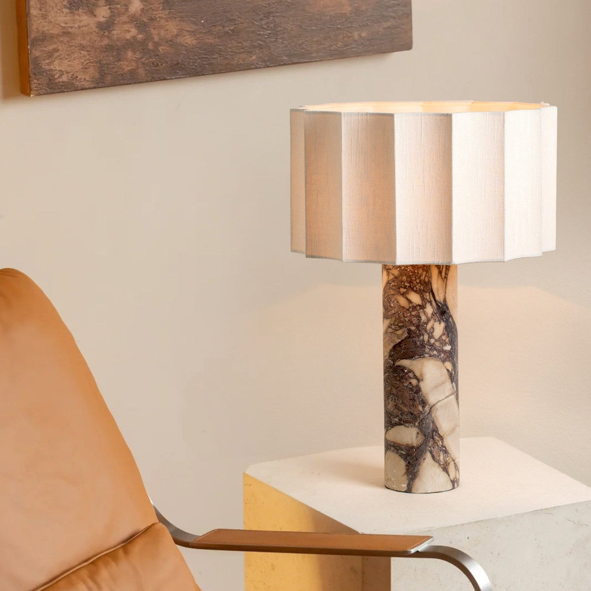 The Clérachic table lamp's faceted cream shade and veined purple marble base stand on a stone plinth beside a tan armchair.