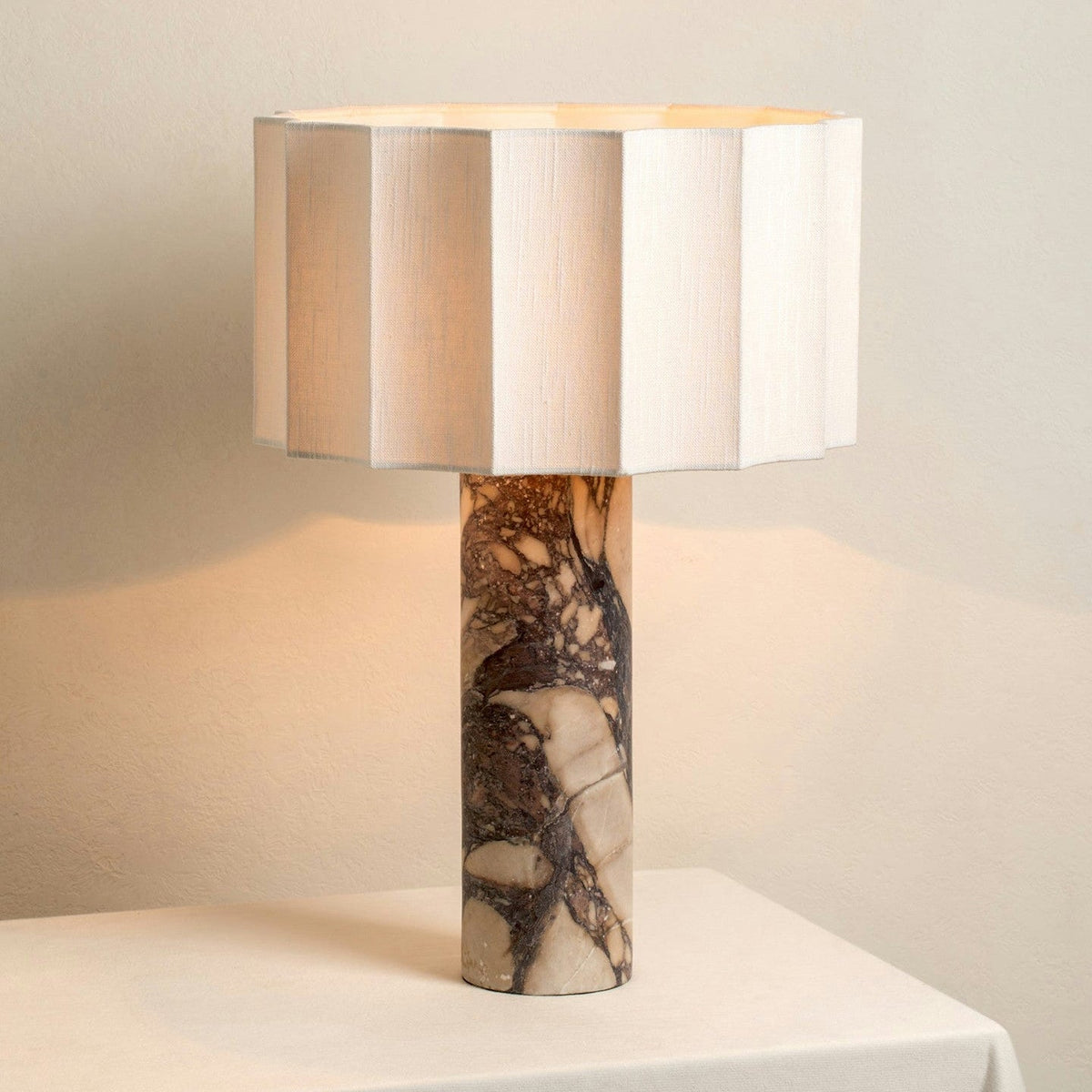 A close view of the Clérachic table lamp's faceted fabric shade glowing warm above its cylindrical purple marble base.