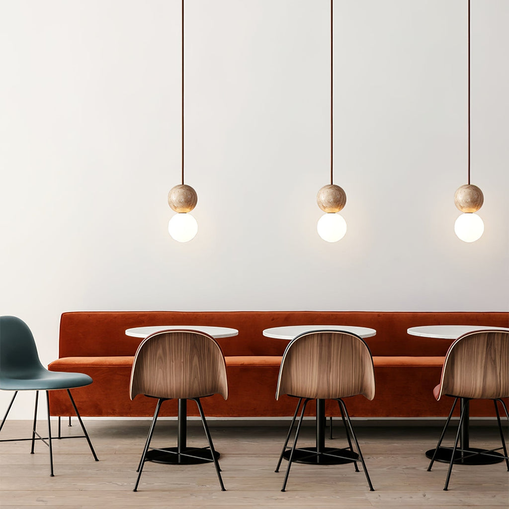 Three staggered Clérachic pendants, travertine spheres over opal glass globes, hang above a rust velvet banquette and wood chairs.