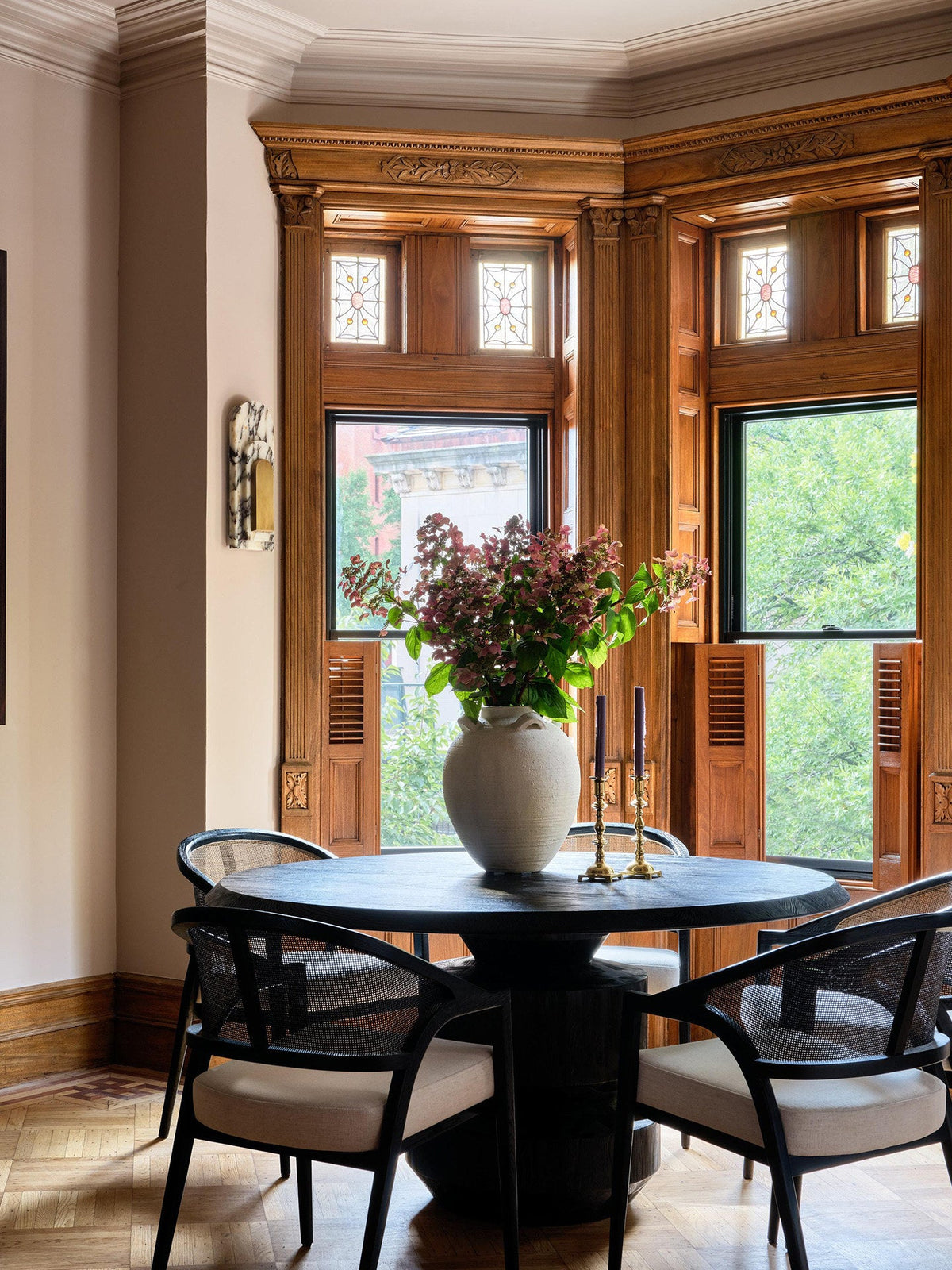 A wider view shows the Clérachic marble sconce mounted on the corner wall of a bay-window dining nook with a round table and cane chairs.