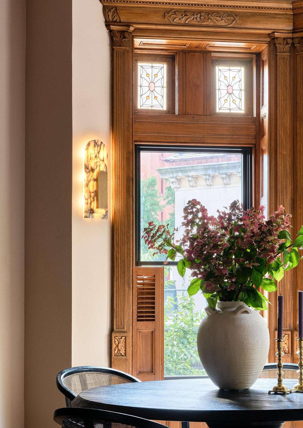 The same Clérachic marble sconce beside the bay window, cropped close, with the flower-filled vase and brass candlesticks on the table.