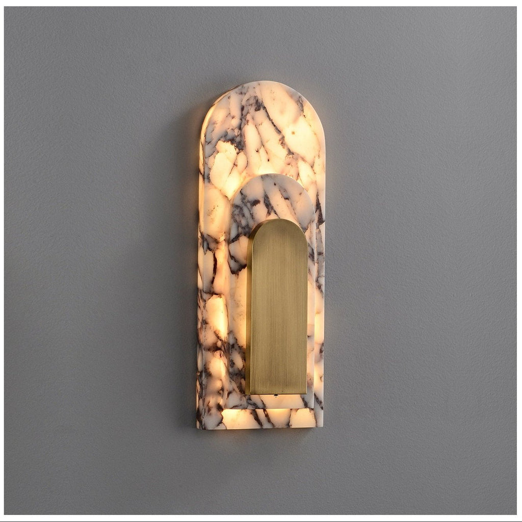 Clérachic marble arch sconce lit against a plain grey wall, light glowing through the veined stone around its brushed brass insert.