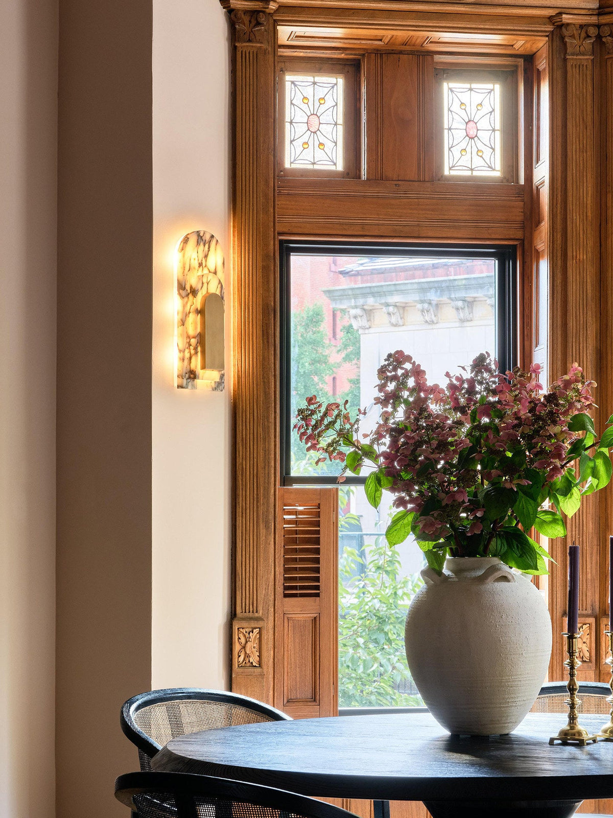 Clérachic marble arch wall sconce glows beside a wood-trimmed bay window, with a vase of pink flowers on the round table below.