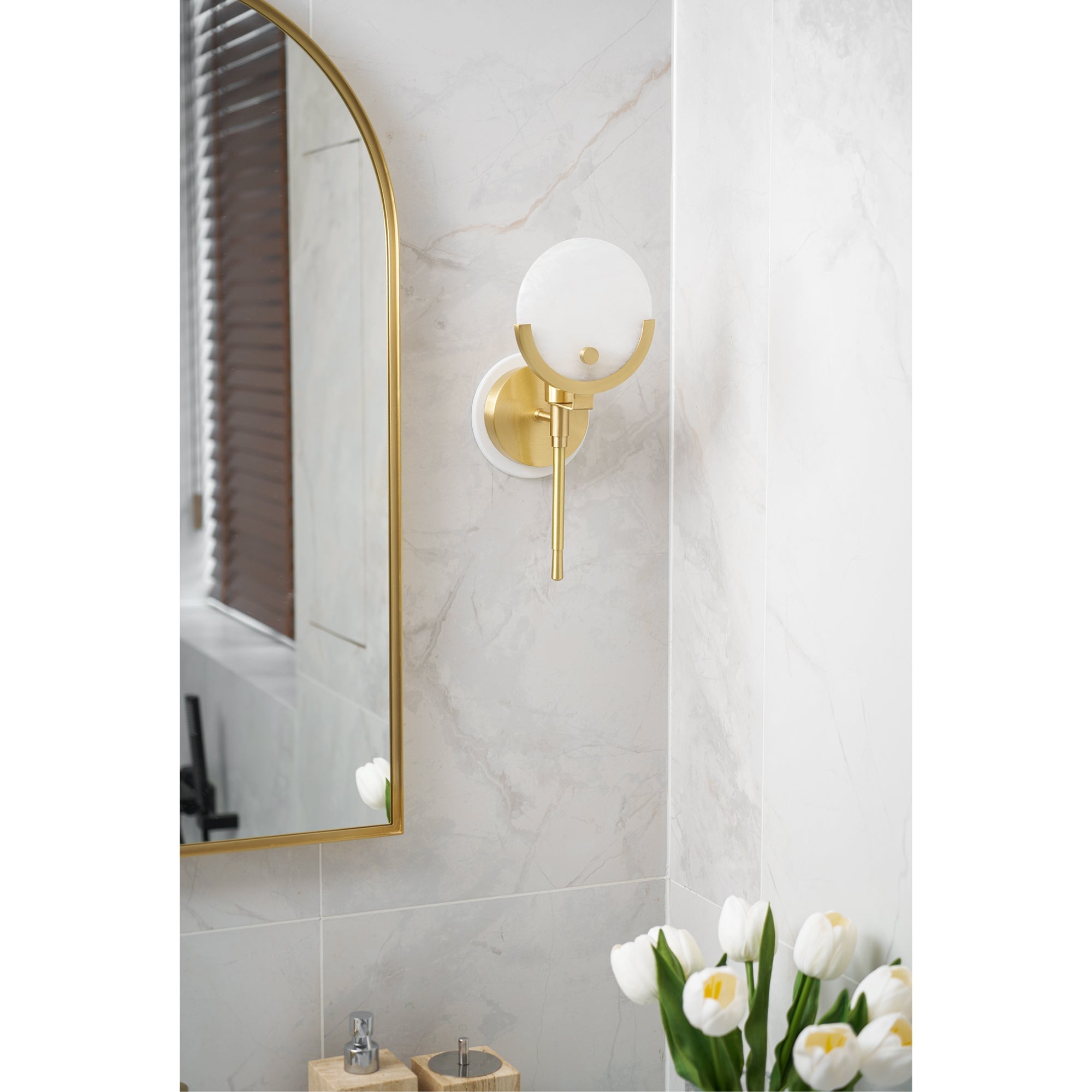 Clérachic zavarara wall light unlit on a marble-tiled bathroom wall next to an arched brass-framed mirror.