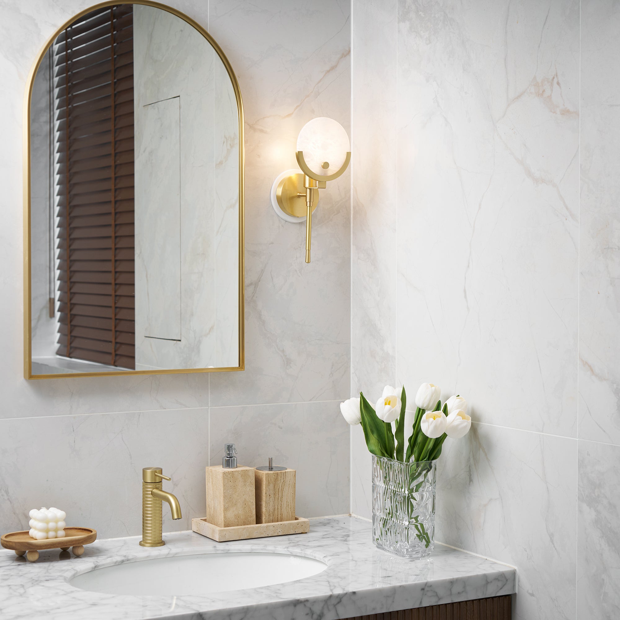 Clérachic wider bathroom view with the Zavarara sconce lit above a marble counter, brass tap and basin below.