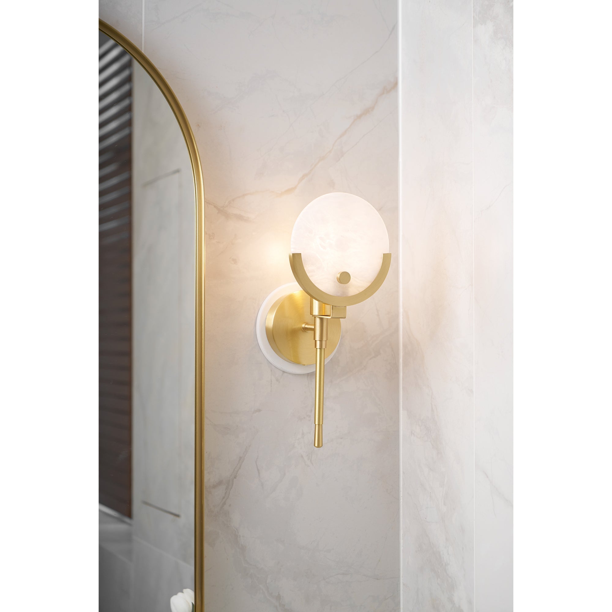 The Clérachic sconce lit on a marble-tiled bathroom wall next to an arched brass-framed mirror.