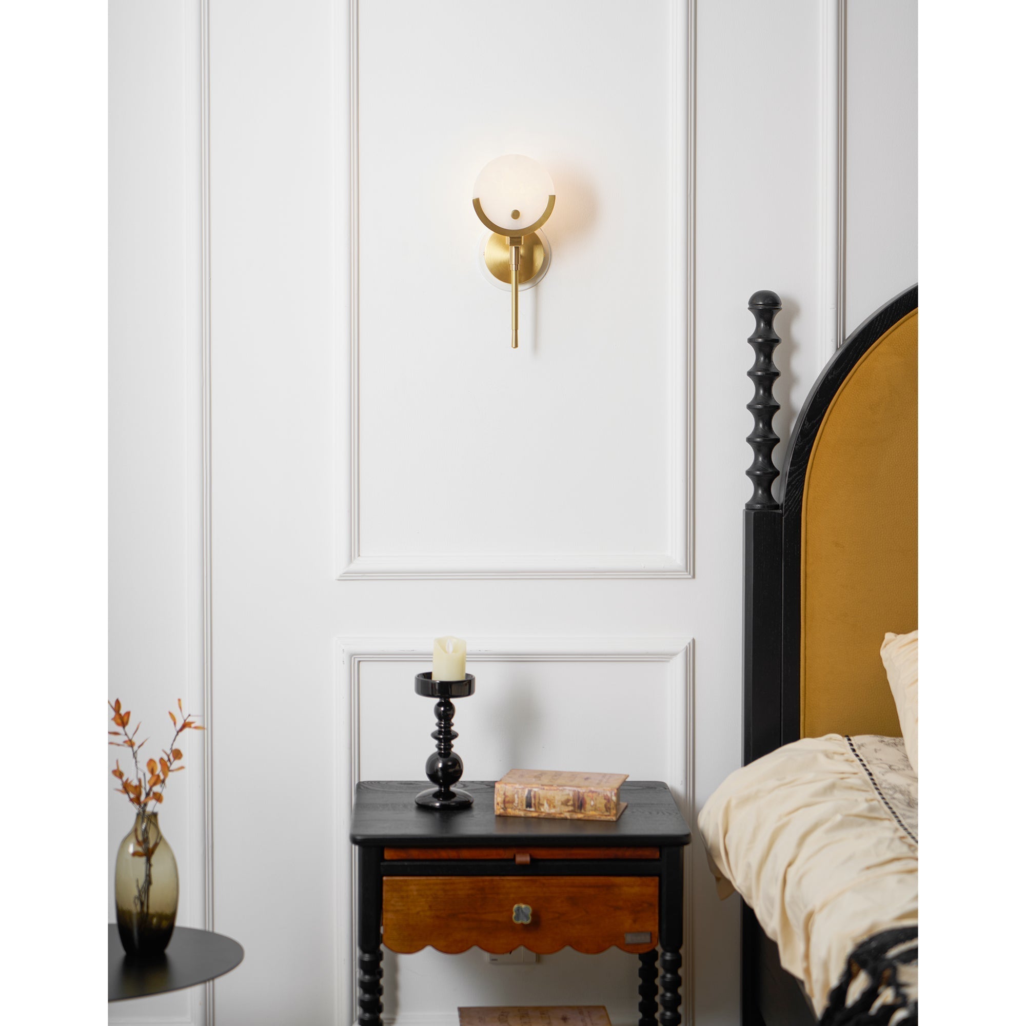 Clérachic compact Zavarara sconce lit on a white panelled bedroom wall beside a black nightstand and candle.