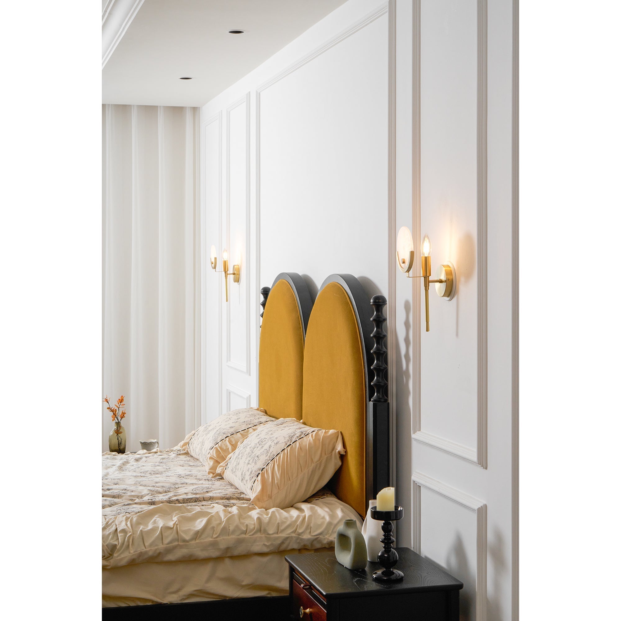 A Clérachic pair of the sconces lit either side of a bed, flanking an arched mustard headboard in a panelled bedroom.
