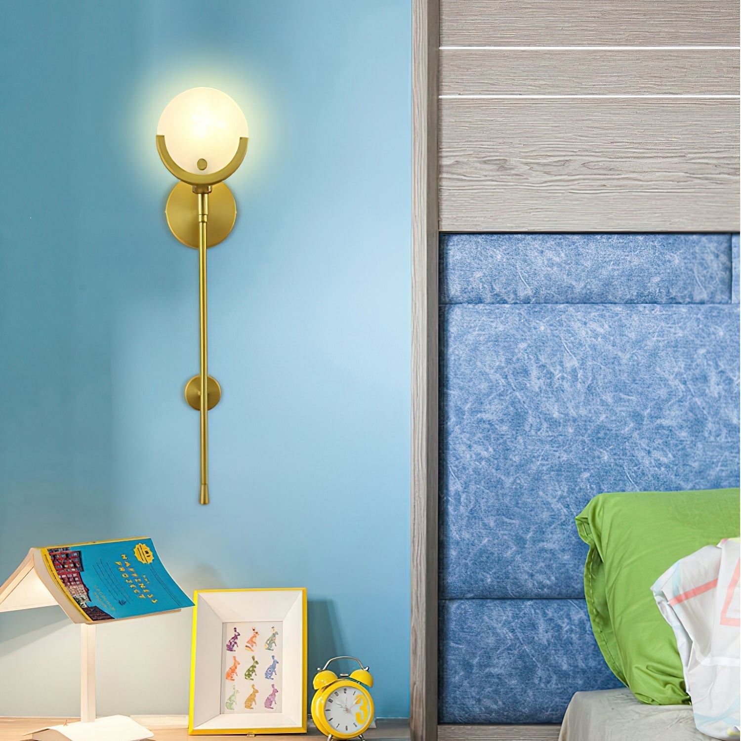 Clérachic tall sconce lit on the blue wall of a child's room above picture frames and a yellow alarm clock.