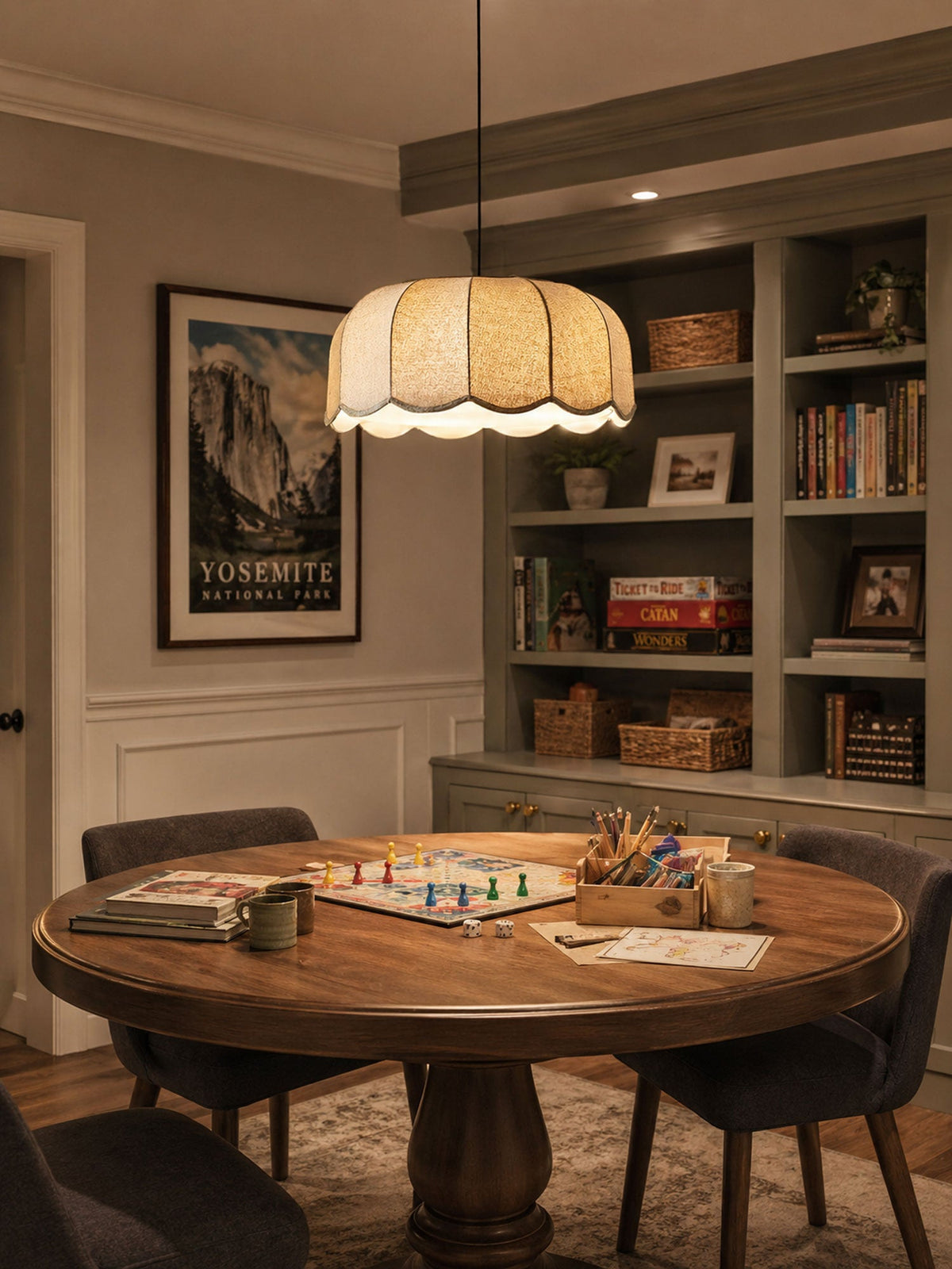 Lit above a round table set for a board game, this beige Clérachic pendant lamp hangs before built-in bookshelves.