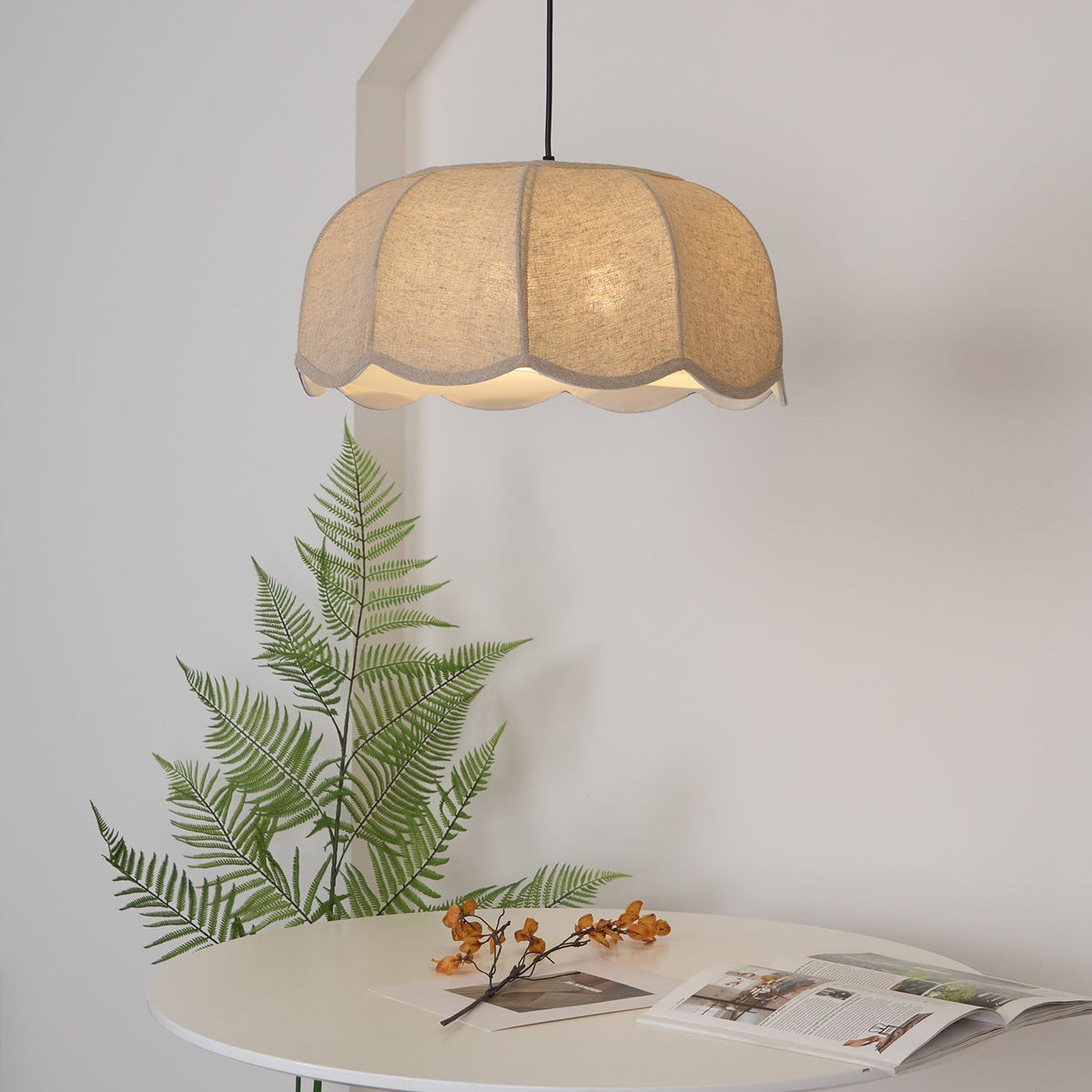 A round white table styled with a fern frond and an open magazine sits beneath this lit beige Clérachic pendant lamp.