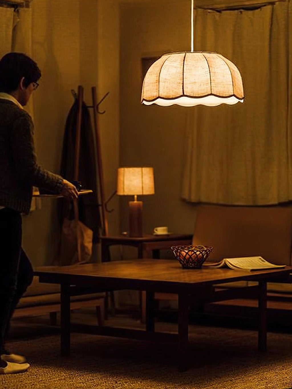 A coat rack and a lit table lamp sit near this beige Clérachic pendant lamp, glowing above a low table.