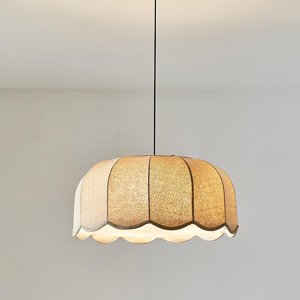 Hanging on a black cord against a plain white wall, this beige Clérachic pendant lamp shows its lit scalloped shade.