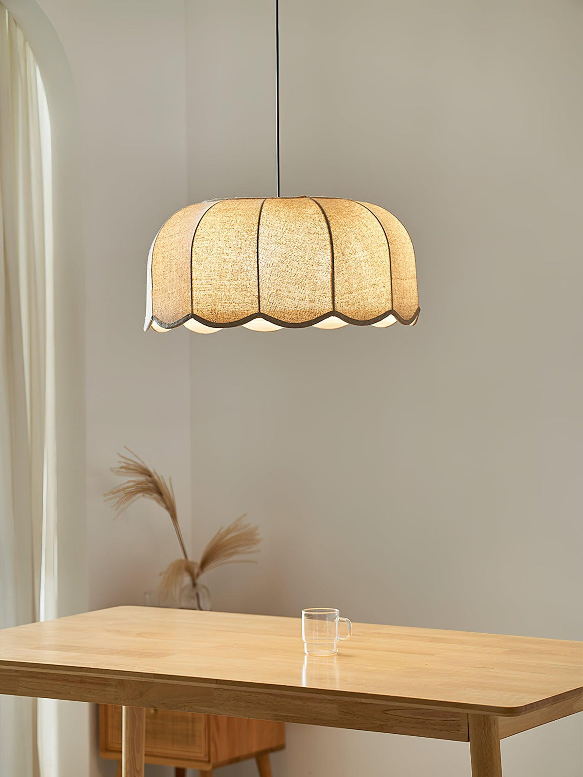 This beige Clérachic pendant lamp hangs above a wooden table set with a glass and a sprig of dried pampas grass.