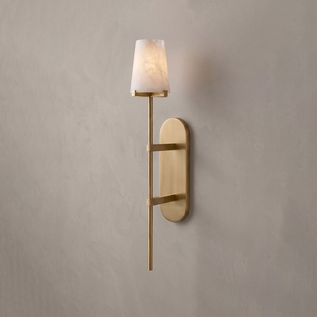 This brushed-brass Clérachic wall sconce glows softly on a plain grey wall, its tall oval backplate catching the light.