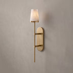 This brushed-brass Clérachic wall sconce glows softly on a plain grey wall, its tall oval backplate catching the light.
