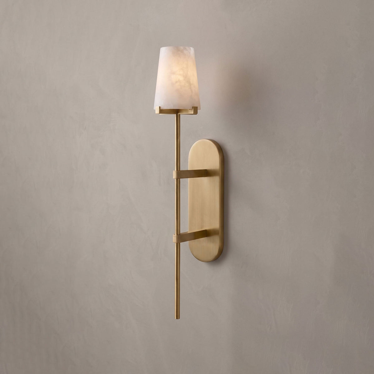 This brushed-brass Clérachic wall sconce glows softly on a plain grey wall, its tall oval backplate catching the light.