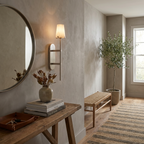 In a hallway, this brass Clérachic wall sconce lights the wall beside a round mirror and a wood bench.