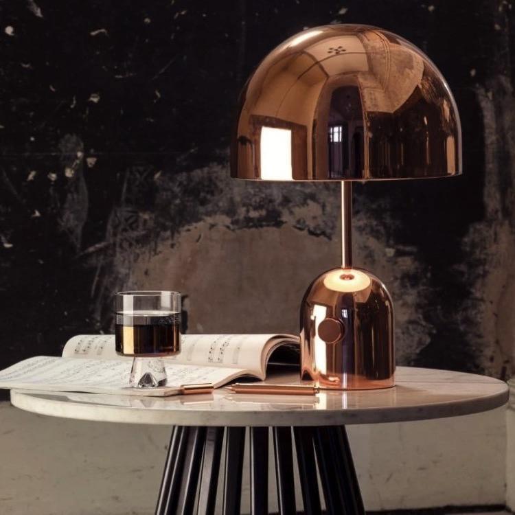 This Clérachic table lamp in rose gold stands lit on a marble table beside a glass and an open magazine.