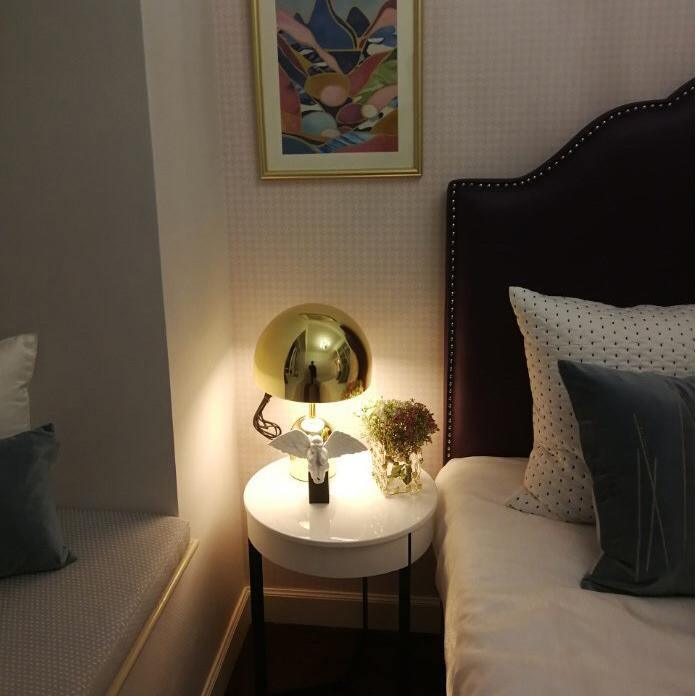 In a bedroom, this Clérachic gold table lamp glows lit on a round white nightstand beside a dark upholstered headboard.