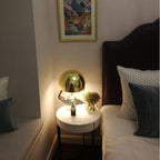 In a bedroom, this Clérachic gold table lamp glows lit on a round white nightstand beside a dark upholstered headboard.