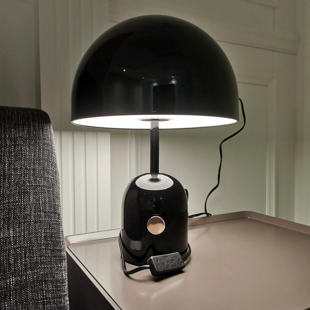 Lit warmly, this Clérachic black table lamp sits on a dark tray table beside a grey armchair, its cord trailing down.