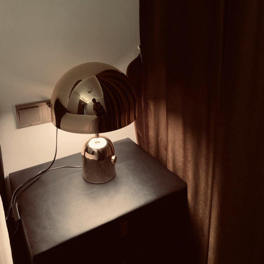 Tucked beside a curtain, this Clérachic chrome table lamp stands on a dark nightstand next to a wall socket.