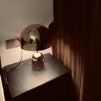 Tucked beside a curtain, this Clérachic chrome table lamp stands on a dark nightstand next to a wall socket.