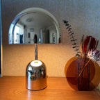 Next to dried flowers and an amber ornament, this Clérachic chrome table lamp stands on a wood table against patterned wallpaper.