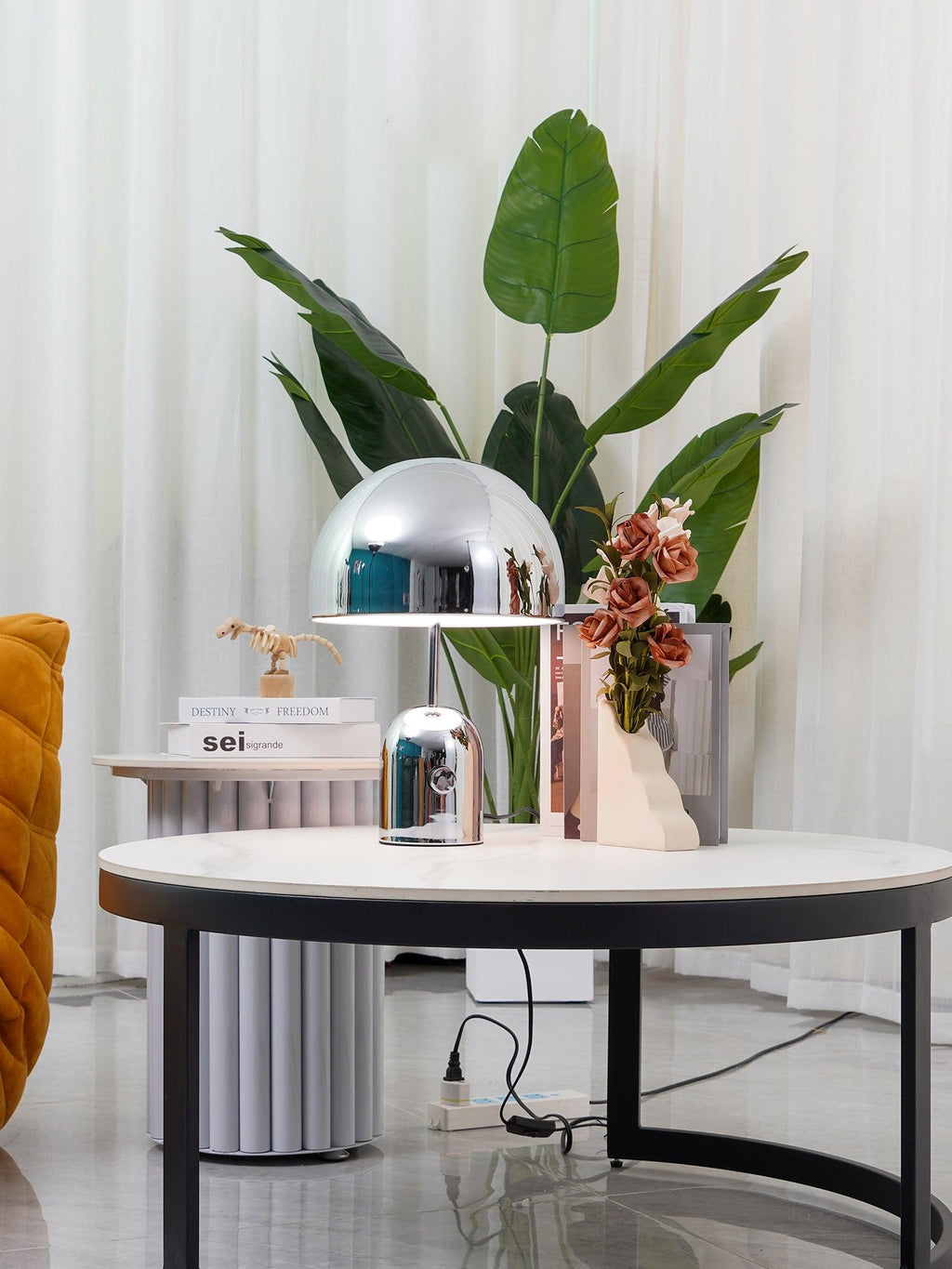 Beside a smaller dome-topped object, this Clérachic chrome table lamp sits on a white round table near potted greenery.