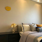 In the corner of a bedroom, this Clérachic gold table lamp sits on a dark nightstand beneath a mustard-yellow clock.