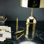 A gold drum-shade lamp stands on dark green marble beside pens and books, a different design from this Clérachic dome lamp.
