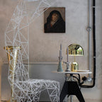 A small dome-topped version of this Clérachic lamp sits on a white pedestal table beside a wire-frame chair and framed portrait.
