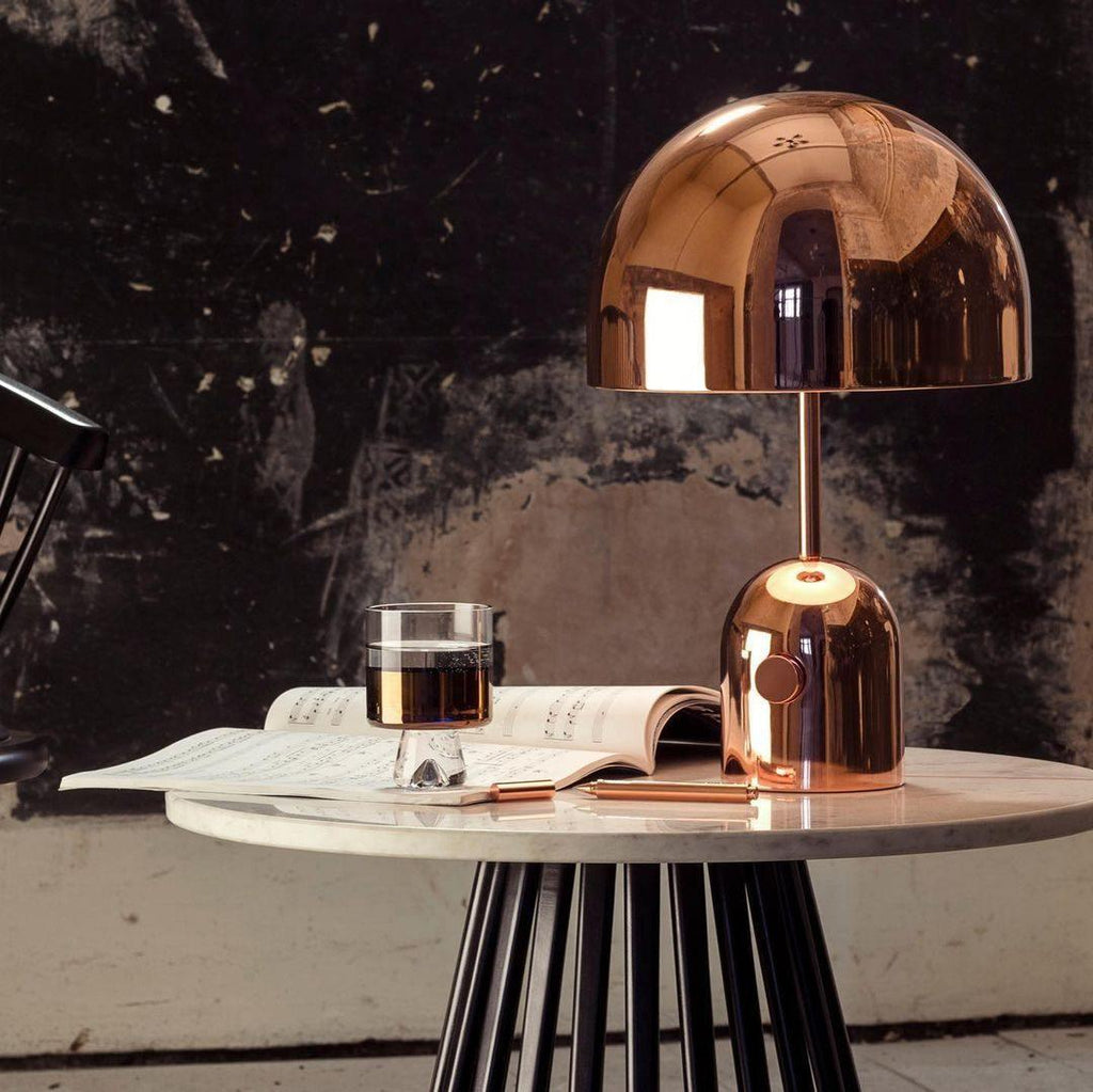 On a round table with a dark textured wall behind, this Clérachic rose gold lamp sits beside a glass and an open magazine.