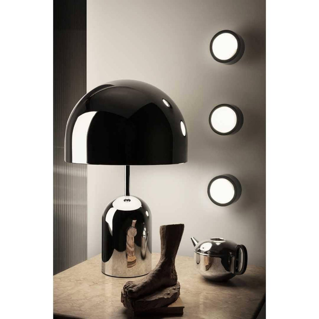 This Clérachic black table lamp stands on a wood table below three round wall sconces, beside a small dark sculpture.