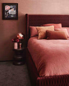 In a bedroom with deep pink velvet bedding, this Clérachic rose gold table lamp stands on a dark round nightstand.