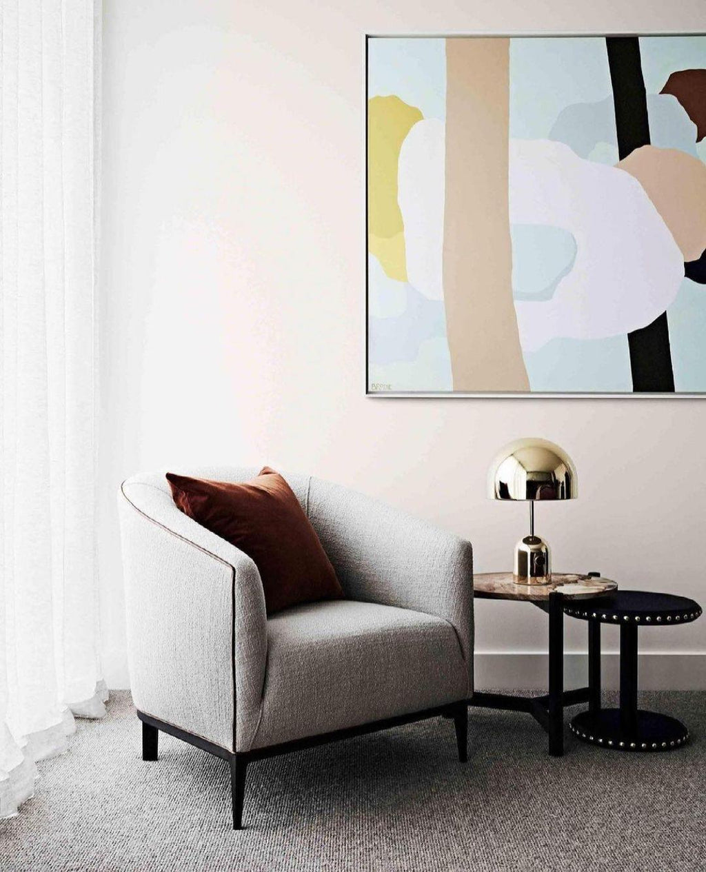 Beside a grey armchair and a pastel abstract painting, this Clérachic gold table lamp sits on a small side table.