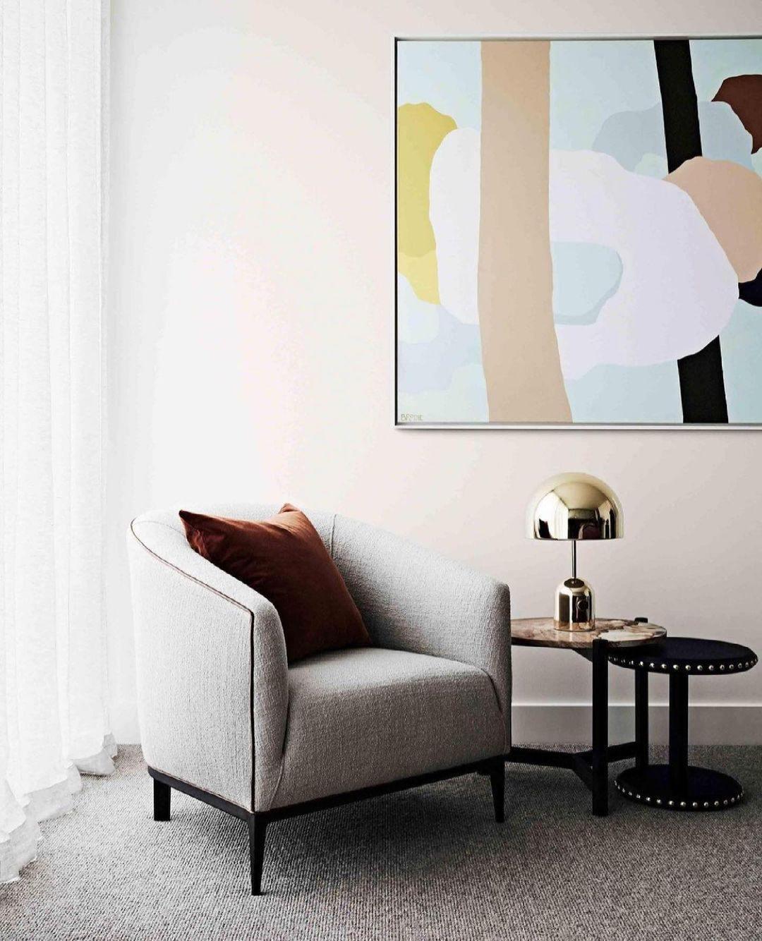 Beside a grey armchair and a pastel abstract painting, this Clérachic gold table lamp sits on a small side table.