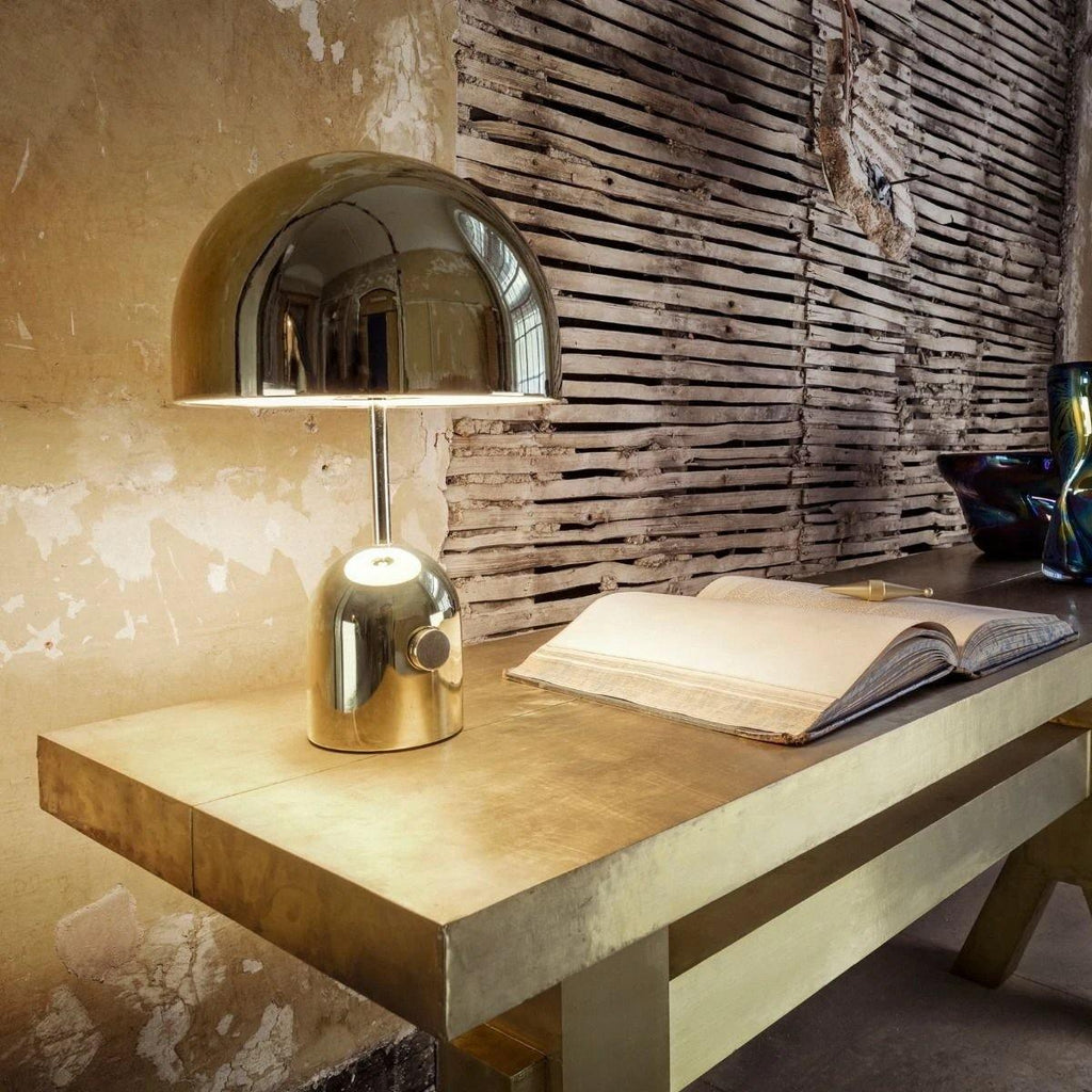 This Clérachic gold table lamp sits on a gilded desk against a distressed plaster wall with an open book nearby.