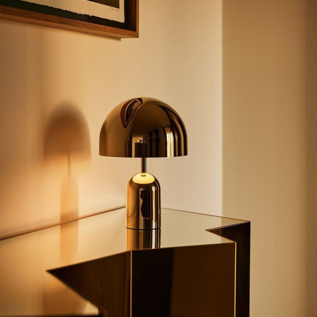 Lit and glowing warm, this Clérachic gold table lamp perches on the corner of a glass table in a softly lit room.
