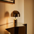 Lit and glowing warm, this Clérachic gold table lamp perches on the corner of a glass table in a softly lit room.