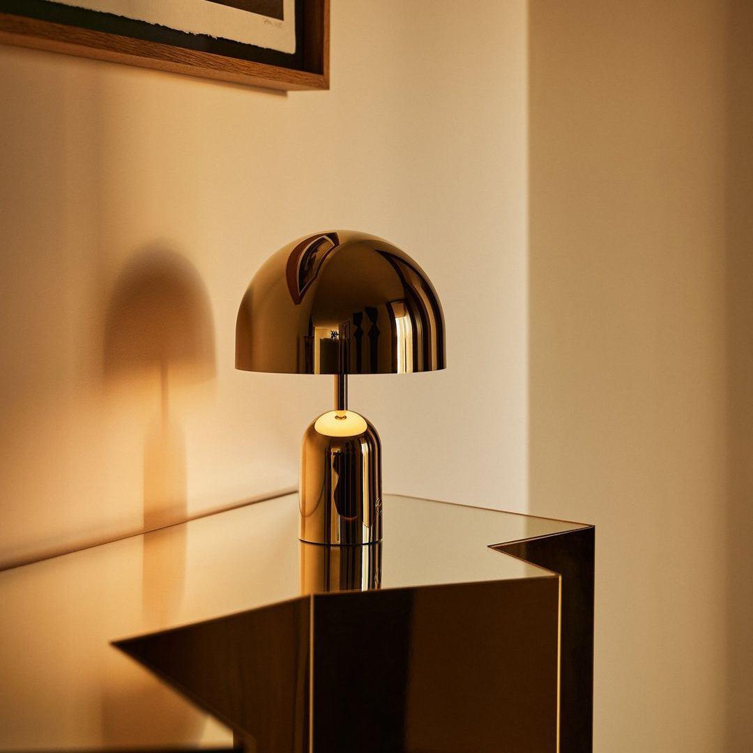 Lit and glowing warm, this Clérachic gold table lamp perches on the corner of a glass table in a softly lit room.